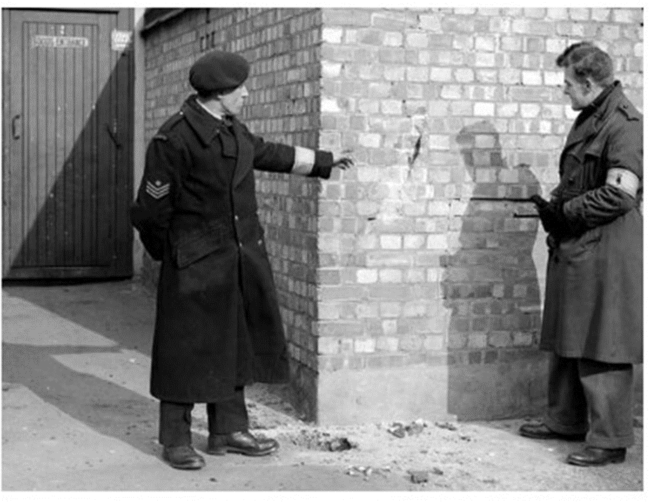 Norfolk based Bomb Reconnaissance Officers examine where an unexploded anti-aircraft shell struck a wall before hitting the ground. (Notice that both men are wearing the Bomb Reconnaissance arm band. The chap on the left is wearing it low on the sleeve, no doubt to avoid covering his other insignia.)