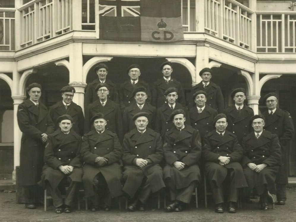 Group of wardens at stand down in May 1945 wearing greatcoats. Note flag and most wearing the CD badge on their berets.